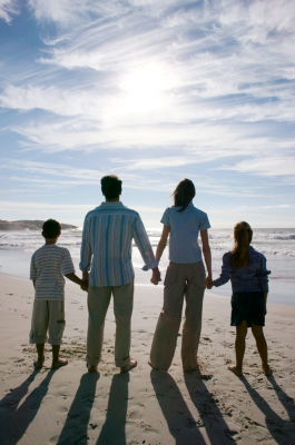 Dad, Mom, and two children holding hands standing on the beach facing the ocean.